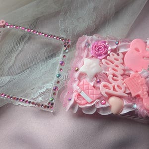 May include: A pink and white phone case with rhinestones and a Barbie logo. The case is decorated with pink and white charms, including a star, a heart, and a comb.