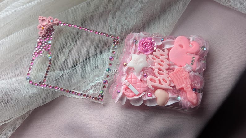 May include: A pink and white phone case with rhinestones and a Barbie logo. The case is decorated with pink and white charms, including a star, a heart, and a comb.