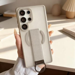 May include: A pale grey phone case with a matching strap and a metal ring. The case features a camera module with multiple lenses. The strap is attached to the back and has a metal ring for holding the phone.