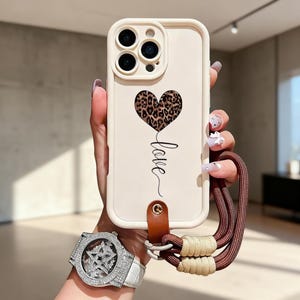 Peut inclure: Coque de téléphone blanc cassé avec un motif de cœur léopard et le mot "love" en cursive. Elle est dotée d'une lanière en cuir marron et d'une dragonne marron et beige. La coque est conçue pour protéger l'appareil photo du téléphone.