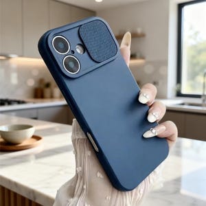 Puede incluir: Una funda de teléfono azul marino con una cubierta para la lente de la cámara. La funda tiene un acabado liso y una cubierta de cámara texturizada. La funda se sostiene en una mano con uñas largas y decoradas. El fondo es una cocina borrosa.
