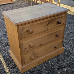 May include: A three-drawer wooden chest of drawers with a light brown finish. It features round wooden knobs on each drawer and a simple, rectangular design. The wood grain is visible. A classic piece of furniture, suitable for a bedroom.