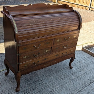 May include: A classic wooden roll-top desk, featuring four drawers and brass handles. The desk has a curved roll-top and elegant, curved legs. The wood has a rich, brown hue. A traditional piece of furniture, likely from a bygone era.