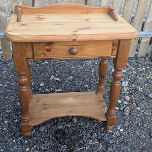 May include: A small, wooden side table with a drawer and a lower shelf. The table is made of light brown wood and has turned legs. The drawer has a round wooden knob. The table's dimensions are approximately 61 cm high, 51 cm wide, and 30 cm deep.