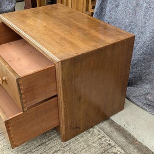May include: A wooden bedside table with two drawers open, showing the inside. The table is a warm brown colour, with visible wood grain. The drawers have small, round wooden knobs. The table is a simple, classic design.