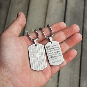 May include: Two silver-coloured metal dog tag pendants on box chains. One tag features an engraved American flag with a central cross. The other tag is engraved with the text: God grant me the Serenity to accept the things I cannot change, Courage to change the things I can, and Wisdom to know the difference. Reinhold Niebuhr.