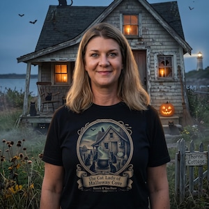 Può includere: Una t-shirt nera a maniche corte con una grafica circolare raffigurante un cottage con gatti neri, un calderone e una zucca. Il testo recita The Cat Lady of Halloway Cove e Knock If You Dare. Il disegno è incorniciato da un bordo a corda.