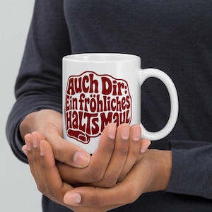 Könnte beinhalten: Ein weißer Keramikbecher mit dunkelrotem Text, der "Auch Dir: Ein fröhliches HALTS MAUL" in einer fetten, stilisierten Schrift zeigt. Der Becher hat eine standardmäßige zylindrische Form mit einem geschwungenen Henkel auf der rechten Seite.