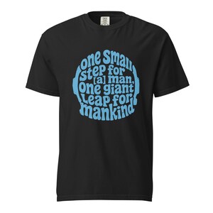 Könnte beinhalten: Schwarzes T-Shirt mit dem Zitat "one small step for [a] man, one giant leap for mankind" in Hellblau. Der Text ist in einem kreisförmigen Design angeordnet. Das Shirt hat einen Rundhalsausschnitt.