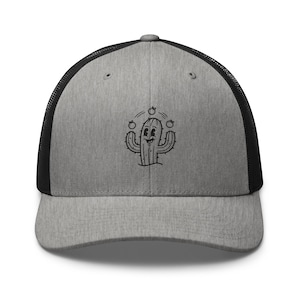 May include: A grey trucker cap with a black mesh back. The front displays a black and white cartoon cactus juggling three small fruits. The cap has a curved peak and a snapback fastening.