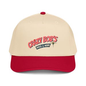 Op de afbeelding: Een crèmekleurige baseballpet met een rode klep en een rode knoop aan de bovenkant. Op de voorkant staat de tekst "CRAZY BOB'S BIKES & MORE" in een gestileerd rood lettertype met een donkerblauwe banner en een kleine gouden ster.