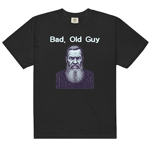 Op de afbeelding: Een zwart t-shirt met korte mouwen en een grafische illustratie van een persoon met een lange baard en een strenge uitdrukking. Boven de illustratie staat de tekst "Bad, Old Guy" in een lichtblauw lettertype. Het shirt heeft een ronde hals.