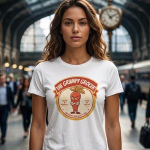 Könnte beinhalten: Ein weißes kurzärmeliges T-Shirt mit einer kreisförmigen Grafik, die eine rote, anthropomorphe Karotte mit verschränkten Armen zeigt. Der Text lautet THE GRUMPY GROCER, FRESH PRODUCE, GOOD PRICES und GROCERIES WITH AN ATTITUDE.
