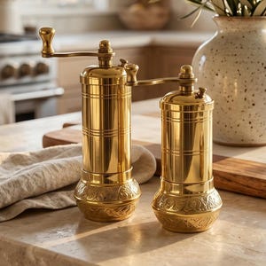 May include: Two cylindrical brass spice grinders with manual hand cranks at the top. Each grinder features a smooth upper body with horizontal etched lines and a wider, flared base decorated with an intricate, embossed floral pattern.