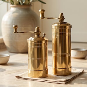 May include: Two cylindrical brass spice grinders with manual hand cranks positioned on a small cloth napkin. The grinders feature a polished metallic finish with horizontal grooved detailing along the body and a small knob on the top handle.