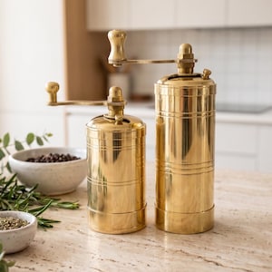 May include: Two cylindrical brass spice grinders with manual hand cranks positioned on top. The grinders feature a polished metallic finish with horizontal grooved detailing along the body. They are accompanied by small speckled ceramic bowls.