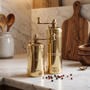 May include: Two brass pepper grinders of different sizes sit on a marble countertop, next to scattered peppercorns. A wooden cutting board, small wooden bowls, and a spoon are in the background. The kitchen setting has a warm, inviting ambiance.