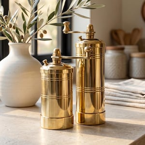 May include: Two cylindrical brass spice grinders with manual hand cranks positioned on top. The grinders feature a polished metallic finish with horizontal grooved detailing around the body. The set includes one shorter and one taller unit.