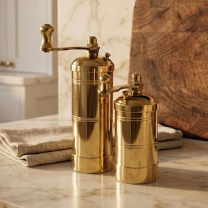 May include: Two brass pepper grinders with a wooden cutting board and a bottle of olive oil. The grinders are different sizes and have a classic design with a crank handle. The olive oil bottle is green with a white label.