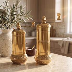 May include: Two cylindrical brass spice grinders with manual hand cranks at the top. Each grinder features a smooth upper body and a base decorated with intricate embossed floral patterns. The metal has a polished gold-toned finish.