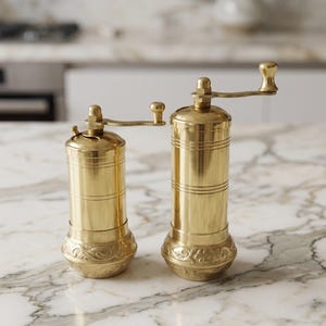 May include: Two cylindrical brass spice grinders of different heights, each featuring a smooth upper body with horizontal line detailing and a base decorated with intricate embossed floral patterns, topped with a small manual crank handle.