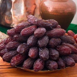 May include: A display of dark brown dates on a decorative plate. Other plates of dates are visible, along with a silver container and a copper-coloured vase. The dates have a wrinkled texture and are arranged in a visually appealing display.