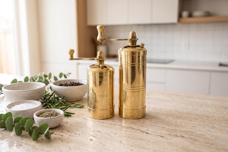 May include: Two cylindrical brass spice grinders with manual hand cranks positioned on top. The grinders feature a polished metallic finish with horizontal grooved detailing along the body. They are accompanied by small speckled ceramic bowls.