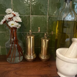 May include: Two cylindrical brass spice grinders with hand cranks, a clear glass vase containing white daisy-like flowers with brown stems, a large clear glass bottle filled with olive oil, and a white marble mortar and pestle set.