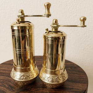 May include: Two cylindrical brass spice grinders with polished gold-toned surfaces. Each mill features a horizontal handle with a rounded knob at the top and a decorative embossed pattern around the base, showcasing a metallic finish throughout.