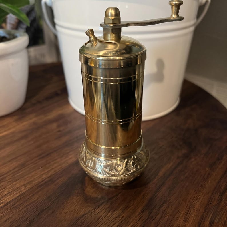 May include: A cylindrical brass pepper mill featuring a polished finish with horizontal line detailing on the body. The base is decorated with an embossed floral pattern, and the top includes a manual crank handle and a small filling knob.