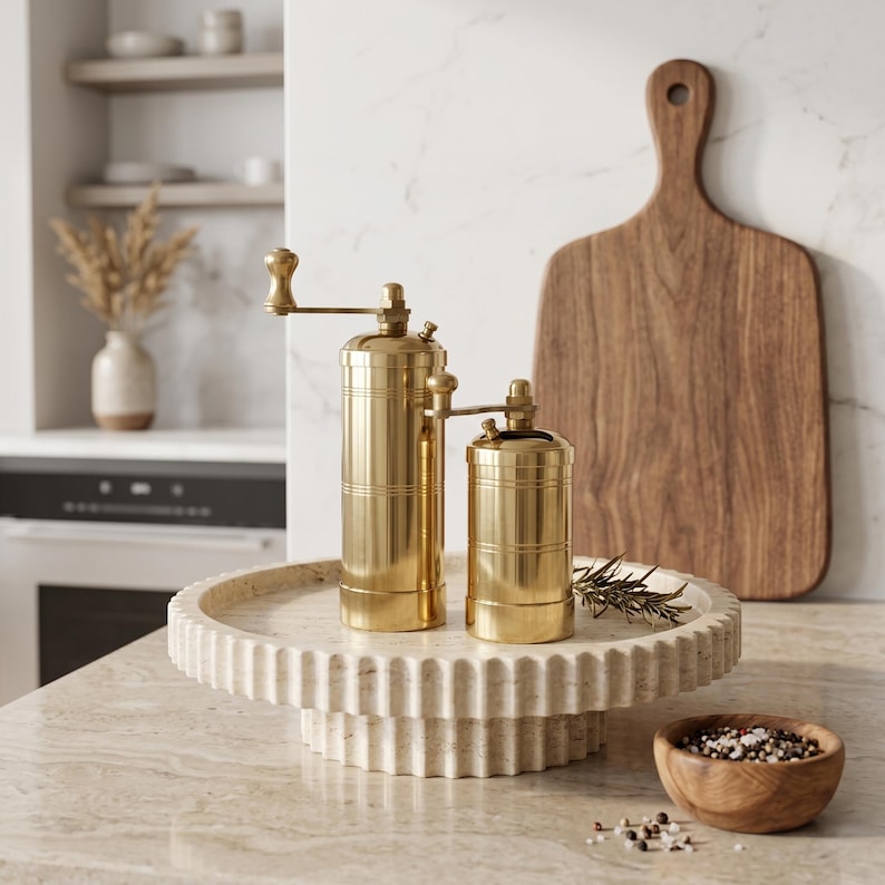 May include: Two cylindrical brass spice grinders with manual hand cranks, featuring a polished metallic finish and horizontal grooved detailing, positioned on a circular fluted stone pedestal tray alongside a small wooden bowl of peppercorns.