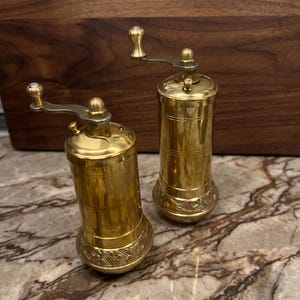 May include: Two cylindrical brass spice grinders with manual hand cranks at the top. Each grinder features a polished metallic finish, a smooth central body with horizontal line detailing, and an ornate floral embossed pattern at the base.