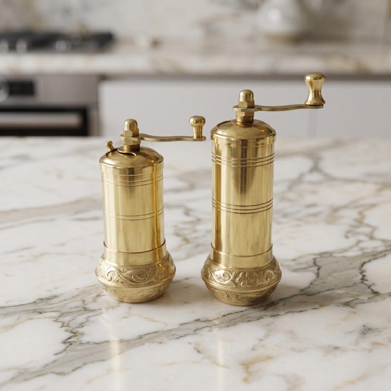 May include: Two cylindrical brass spice grinders of different heights, each featuring a smooth upper body with horizontal line detailing and a base decorated with intricate embossed floral patterns, topped with a small manual crank handle.