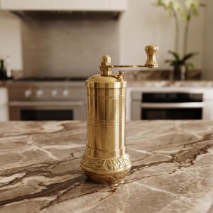 May include: A cylindrical brass pepper mill featuring a smooth upper body and a base decorated with intricate floral and scroll engravings. The mill includes a top-mounted horizontal handle with a small knob for manual grinding operation.