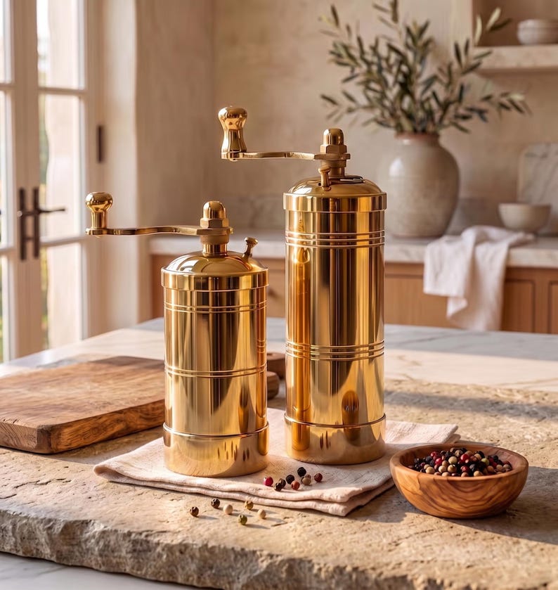 May include: Two cylindrical gold-colored metal spice grinders with manual hand cranks on top. The grinders feature a polished, reflective finish with horizontal grooved detailing around the body. They are positioned upright on a light-colored cloth.