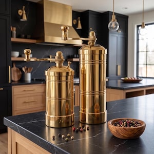 May include: Two cylindrical brass spice grinders with top-mounted hand cranks. The grinders feature a polished metallic finish and are positioned on a dark surface alongside a small wooden bowl containing a mix of whole peppercorns.