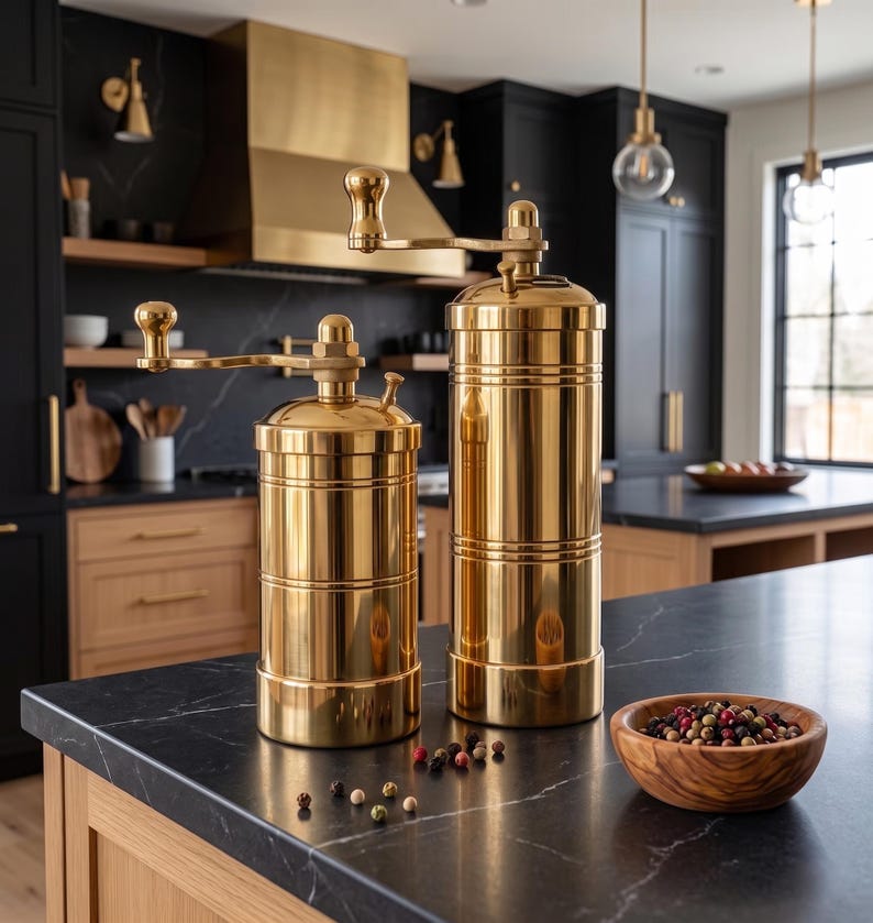 May include: Two cylindrical brass spice grinders with top-mounted hand cranks. The grinders feature a polished metallic finish and are positioned on a dark surface alongside a small wooden bowl containing a mix of whole peppercorns.