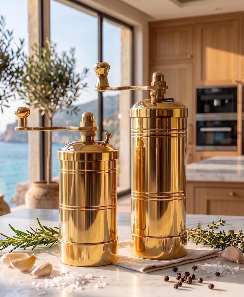 May include: Two cylindrical gold-toned metal spice grinders with manual hand cranks on top. The grinders feature a polished, reflective finish with horizontal grooved detailing around the body. They are positioned upright on a flat surface.