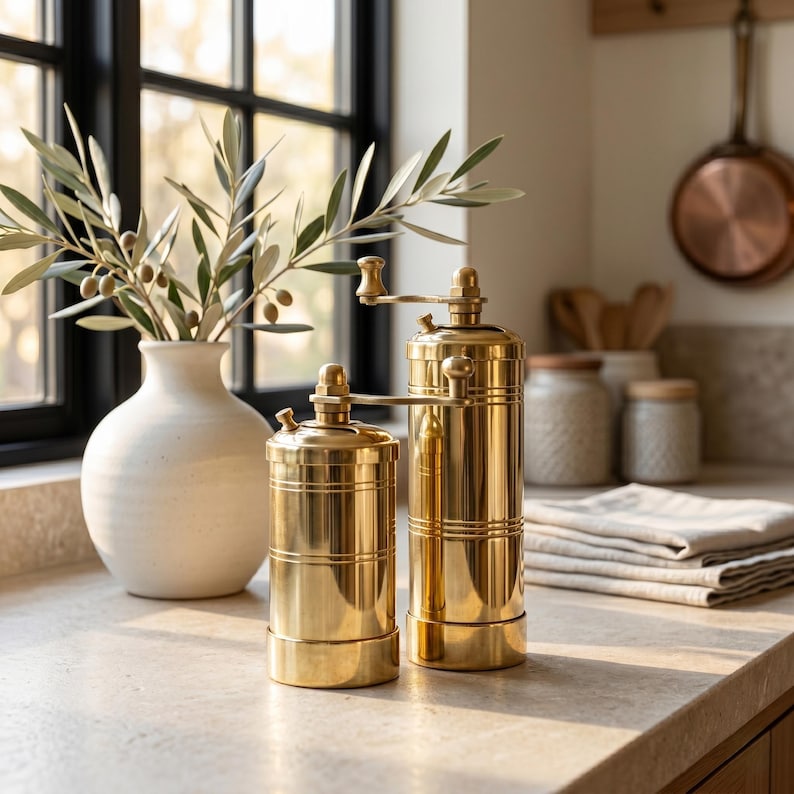 May include: Two cylindrical brass spice grinders with manual hand cranks positioned on top. The grinders feature a polished metallic finish with horizontal grooved detailing around the body. The set includes one shorter and one taller unit.