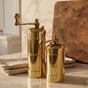 May include: Two cylindrical spice grinders made of polished gold-toned metal. Each grinder features a vertical body with horizontal grooved lines, a top-mounted manual crank handle with a rounded knob, and a small adjustment nut on the top lid.
