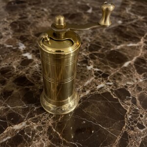 May include: A cylindrical brass pepper mill featuring a polished metallic finish with horizontal engraved lines along the body and a decorative embossed pattern at the base. It includes a top-mounted manual crank handle with a rounded knob.