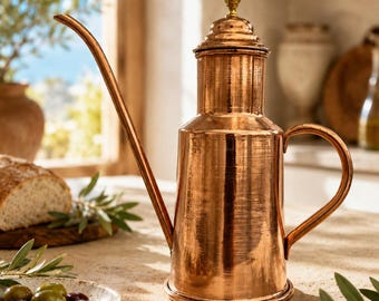 May include: A polished copper oil cruet featuring a tall, slender, curved spout, a cylindrical body with a brushed metallic finish, a matching lid with a small brass-colored knob, and a curved handle attached to the side of the vessel.