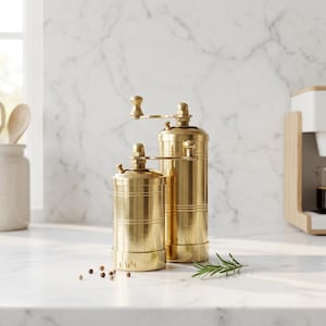 May include: Two cylindrical brass spice grinders with manual hand cranks on top. The grinders feature a polished metallic finish with horizontal grooved detailing around the body. A small sprig of green rosemary and several peppercorns sit nearby.