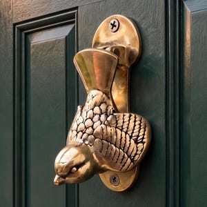 Puede incluir: Un llamador de puerta de latón pulido con forma de pájaro, con plumas de ala detalladas y una cabeza lisa. El pájaro de metal está montado verticalmente en una puerta verde oscuro con dos tornillos visibles en la parte superior e inferior.