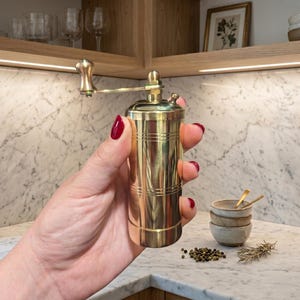May include: A cylindrical brass pepper mill featuring a polished metallic finish, a top-mounted manual crank handle with a rounded knob, and horizontal decorative grooves etched into the body for a classic, functional kitchen tool design.