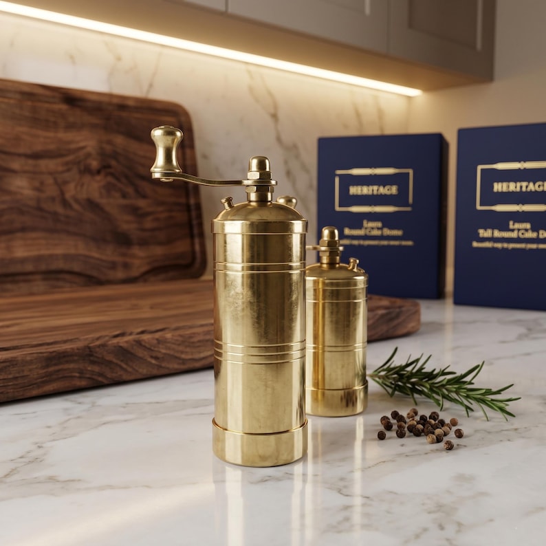 May include: Two cylindrical brass spice grinders with manual hand cranks on top. The taller grinder features a prominent crank handle, while the shorter one sits beside it. A small sprig of rosemary and several peppercorns lie on the surface.