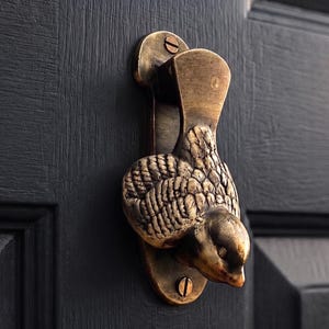 Op de afbeelding: Een koperen deurklopper in de vorm van een vogel met gedetailleerde vleugelveren en een gladde kop. Het metalen beslag is verticaal bevestigd met twee zichtbare schroeven op een donkere, getextureerde deur en heeft een verouderde afwerking.
