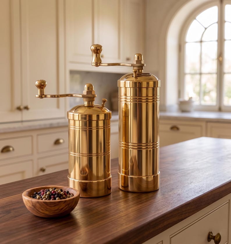 May include: Two cylindrical brass spice grinders with manual hand cranks positioned on top. The grinders feature a polished metallic finish with horizontal grooved detailing. A small wooden bowl filled with mixed peppercorns sits nearby.