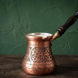 Könnte beinhalten: Eine Kaffeekanne aus Kupfer mit einem komplizierten eingravierten Blumenmuster auf dem Körper, einem breiten Rand mit einer kleinen Ausgießtülle und einem langen, dunkelbraunen Holzgriff, der mit einem messingfarbenen Metallanschluss befestigt ist.