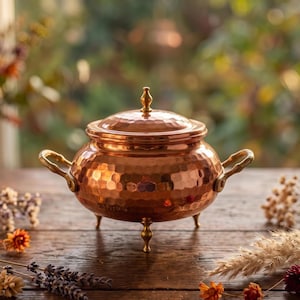 May include: A round, hammered copper pot with a matching lid and a small finial handle. The pot features two curved brass side handles and stands on three slender brass legs. The exterior surface has a reflective, textured metallic finish.